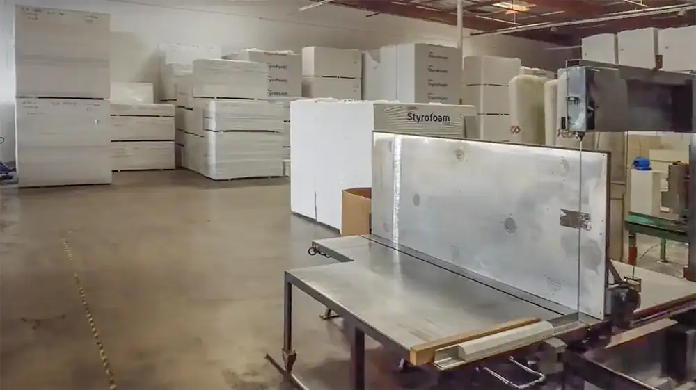A foam cutting machine is seen in a manufacturing warehouse, surrounded by stacks of Styrofoam and other packaging materials.