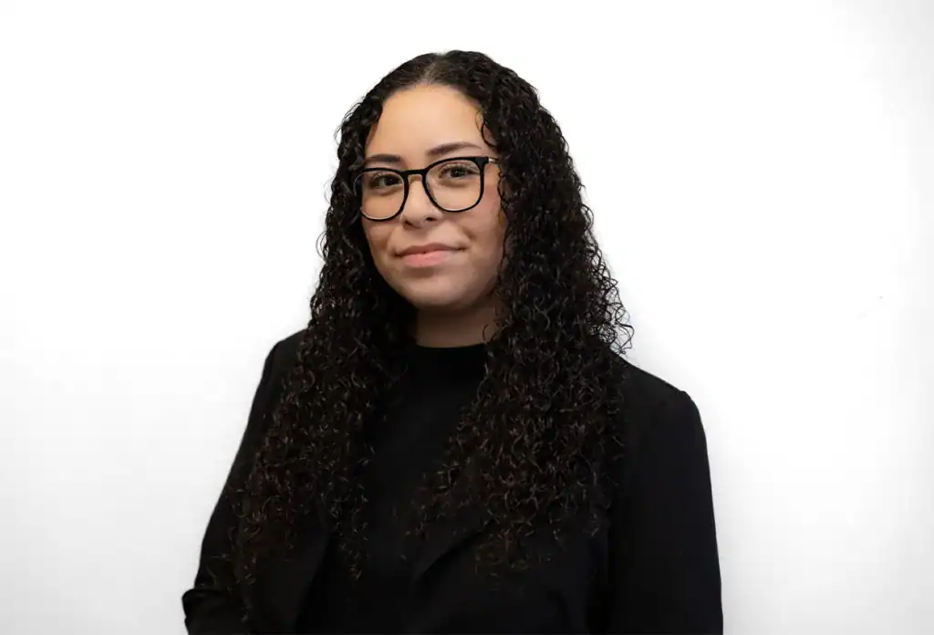 A confident woman with curly hair and glasses, wearing a black blazer, standing against a plain white background.