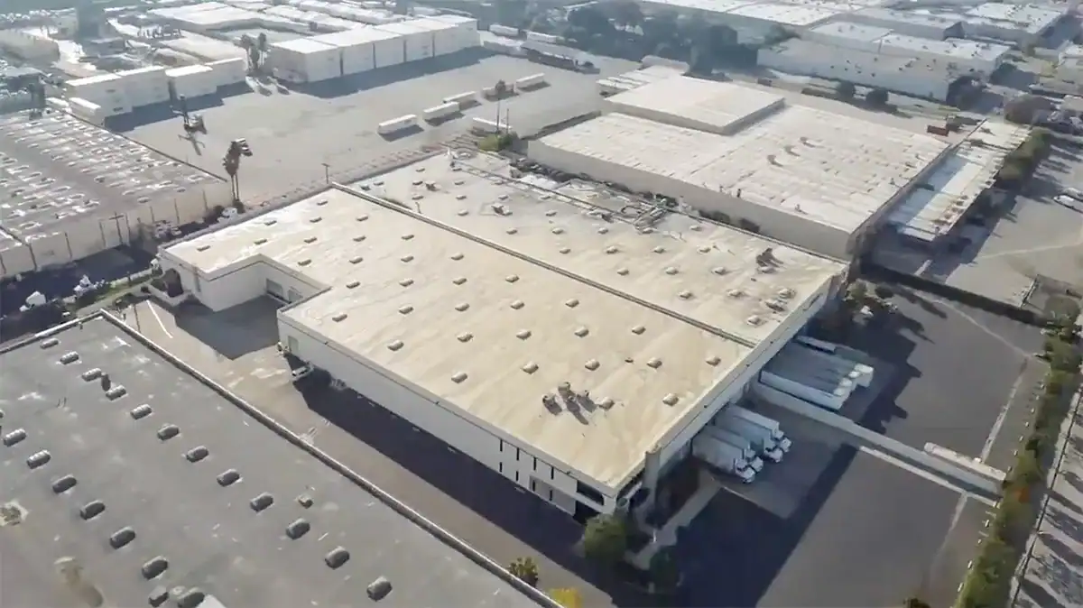 Aerial shot of a large industrial warehouse with a flat white roof and surrounding parking lot.