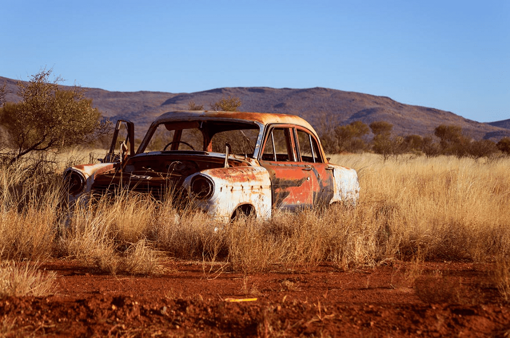 Old rusty car abandoned in dry grassland with mountains in background.