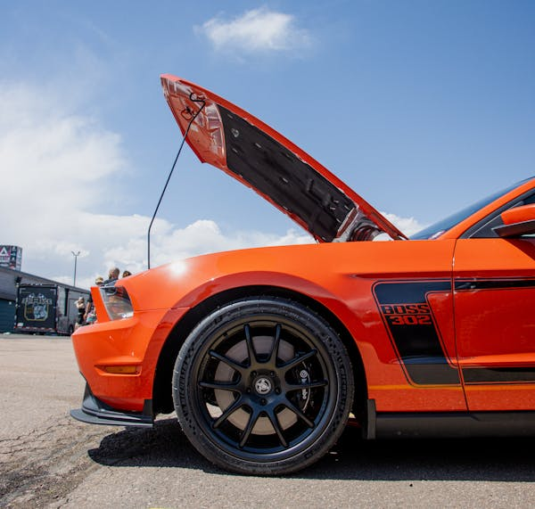 Close-up of a bright orange hot rod with custom black wheels and open hood at a car show.