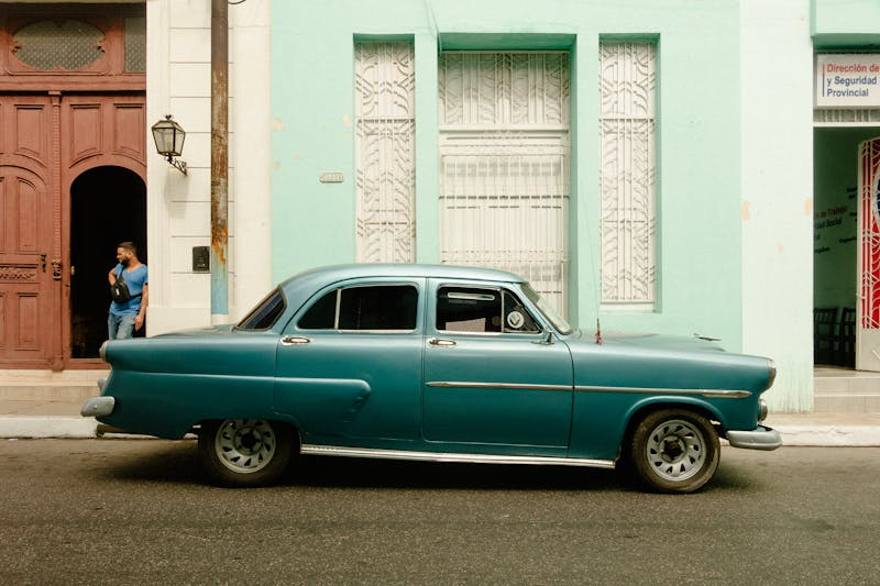 Vintage blue classic car parked on city street with pastel buildings in the background.