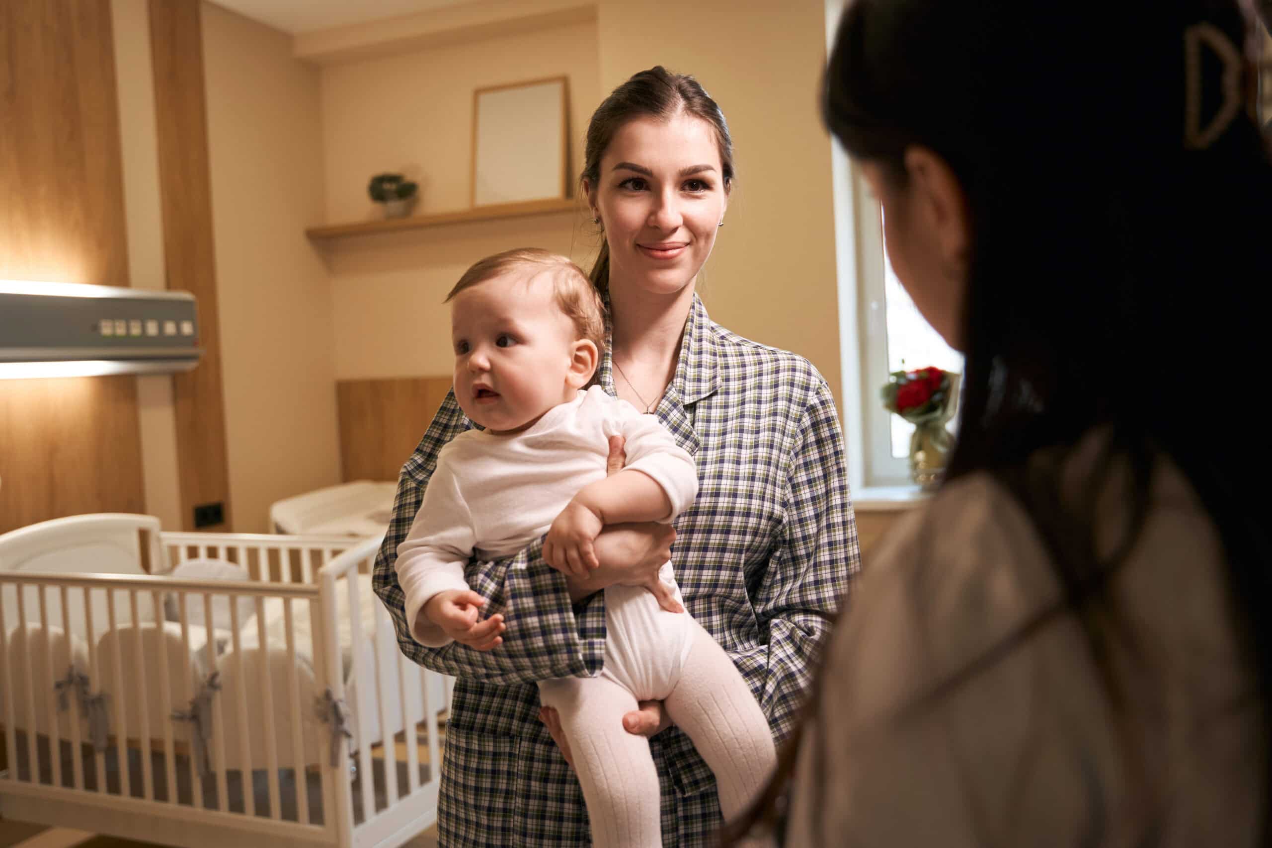 Baby caregiver smiling and holding infant towards a mom while in nursery