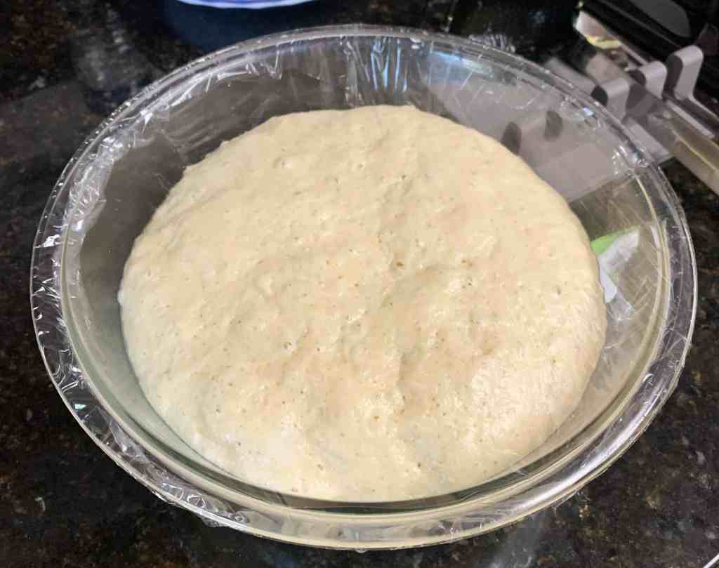 Soft dough rising in a clear bowl covered with plastic wrap, ready for baking or cooking.
