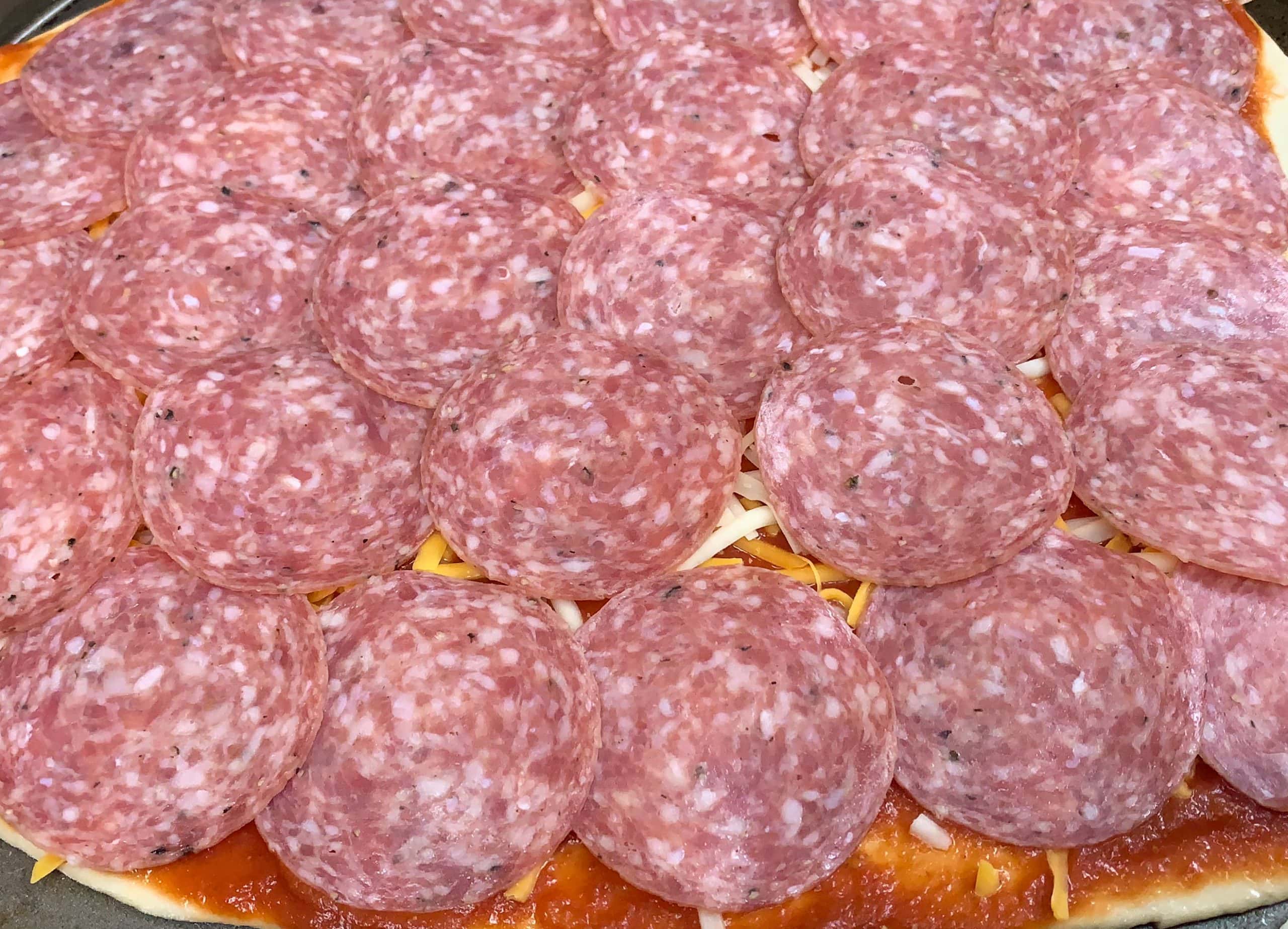 Thinly sliced cured salami on pizza with cheese and tomato sauce, ready to bake.