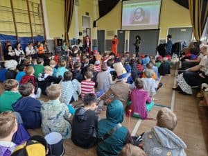 A large group of children and adults attending a presentation or performance in a spacious hall.