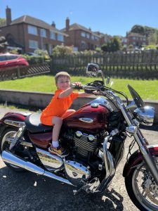 Child enjoying a ride on a motorcycle at Inchmarlo estate. Scenic outdoor setting with residential buildings in the background.