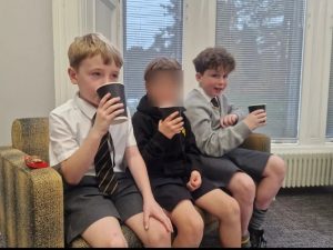 Three children sitting on a sofa, holding cups, smiling, in a classroom setting.