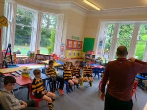 Bright classroom with young children and teacher engaged in activity, large windows, colorful decor.