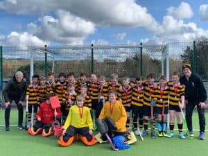 Group of young rugby players in striped jerseys on a sports field, with coaches and a goalpost in the background.