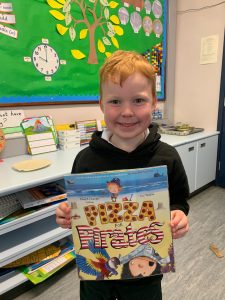 Happy boy holding "Maza Stories" book in classroom.