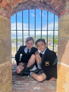 Two children sitting by a castle window with a scenic view, wearing school uniforms.
