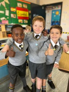 Three happy schoolchildren giving thumbs up in classroom.