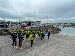 Group of people in bright jackets walking along a harbor shoreline with boats and cloudy sky.