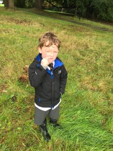 Young boy standing outdoors on lush green grass in a scenic park setting.
