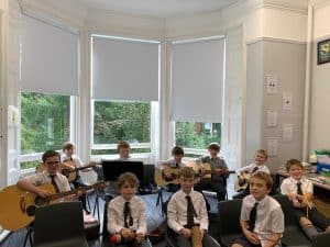 Children playing guitar in classroom with large windows, bright natural light, and cheerful atmosphere.
