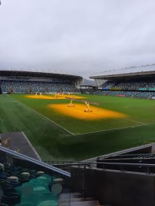 A wide view of Inchmarlo cricket ground with lush green pitch and enthusiastic stadium seating under a cloudy sky.