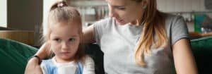 Sad young girl with an upset expression sitting next to a woman, possibly a caregiver or teacher, in a classroom or learning environment.