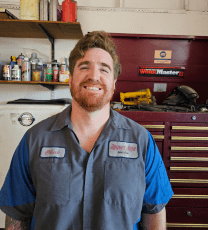 Auto repair technician smiling in workshop with tools and car parts.