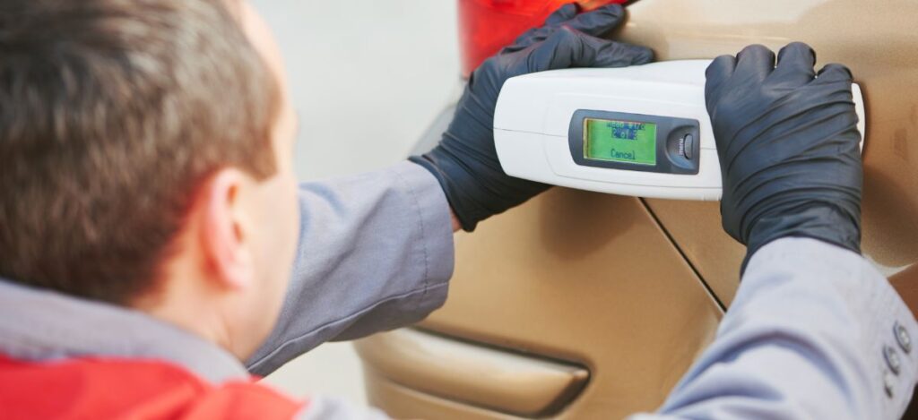 Automotive body repair technician inspecting vehicle with digital paint thickness gauge.