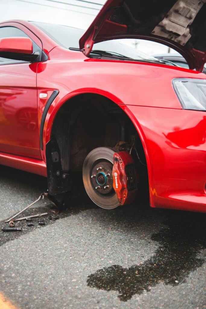 Red car with open hood and missing front wheel, showing brake system.