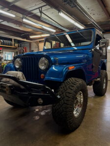 Blue classic Jeep in workshop with large tires and restoration equipment.