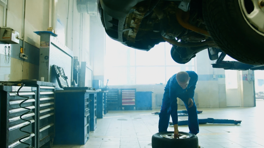 Technician inspecting tire beneath vehicle in auto repair shop.