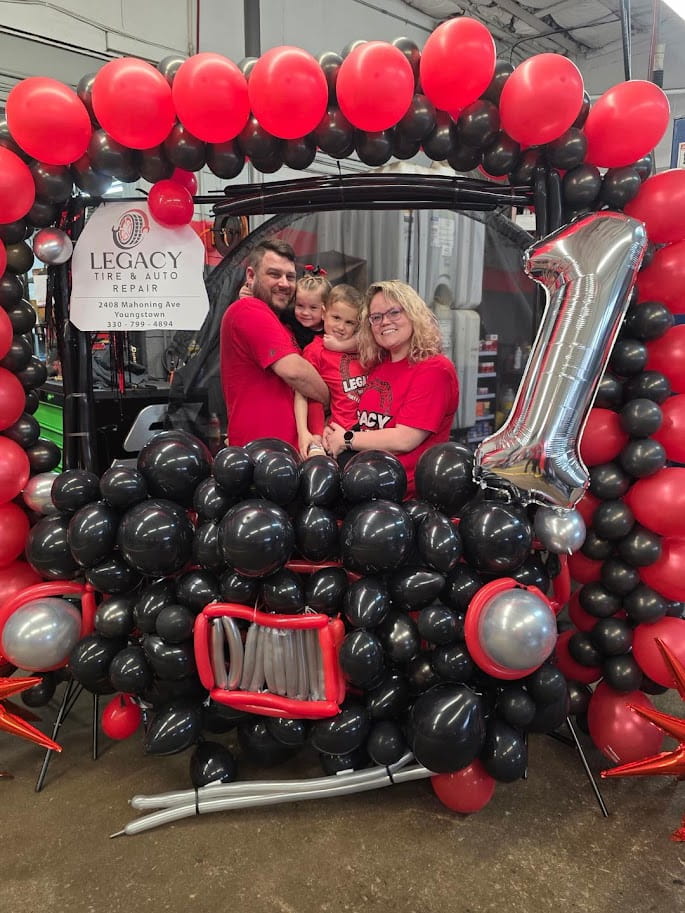 Family celebrating at Legacy Tire & Auto Repair with balloon decorations and a large number one ball.