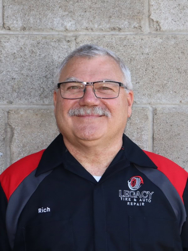 Smiling male employee in Legacy Tire uniform with glasses in front of a brick wall.
