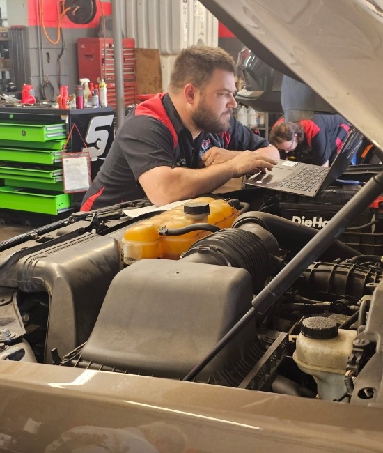 Auto repair technician working on vehicle engine with laptop in garage.