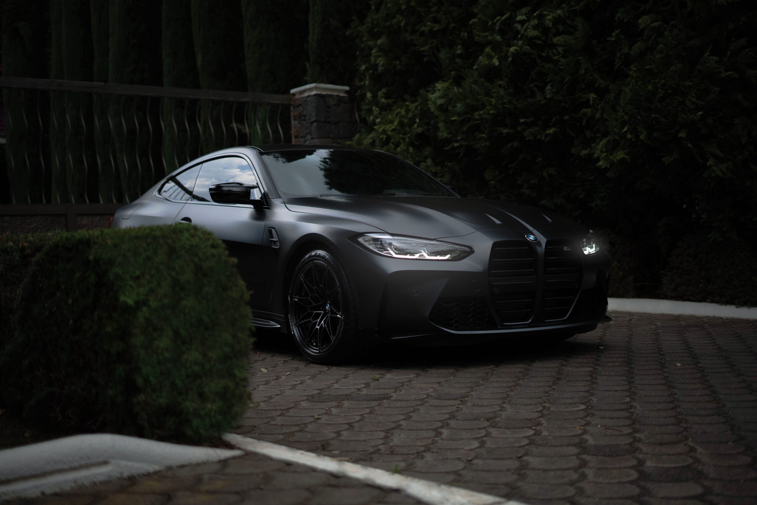 Sleek black BMW M4 parked on a brick driveway with greenery in the background.