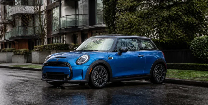 Mini Cooper S blue car parked on wet city street with modern buildings in background.