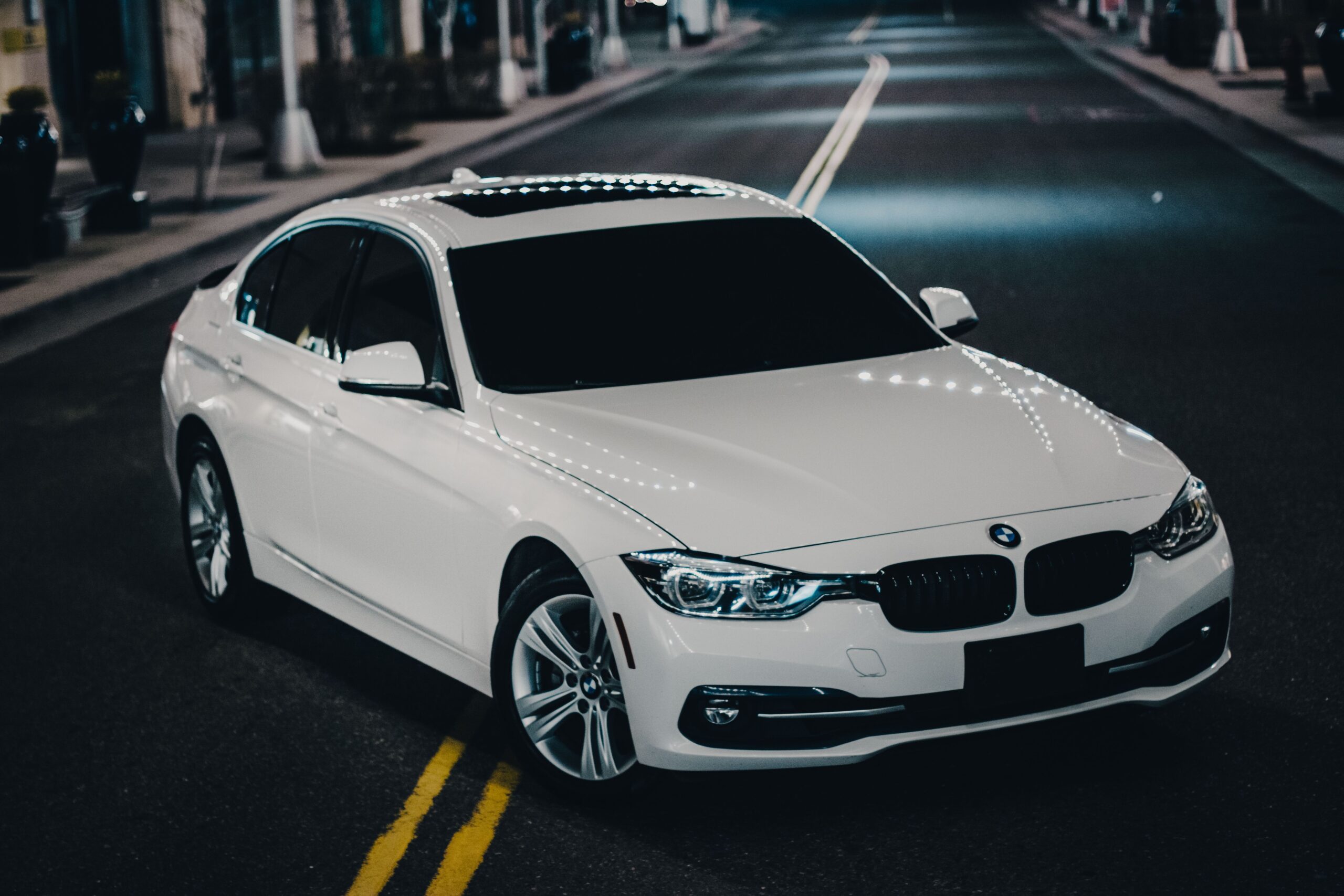 White BMW sedan parked on city street at night with streetlights.