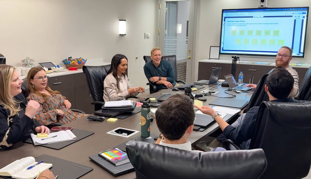 Eight people sit around a conference table in an office, engaged in a meeting while viewing a presentation on a large screen at the front of the room.