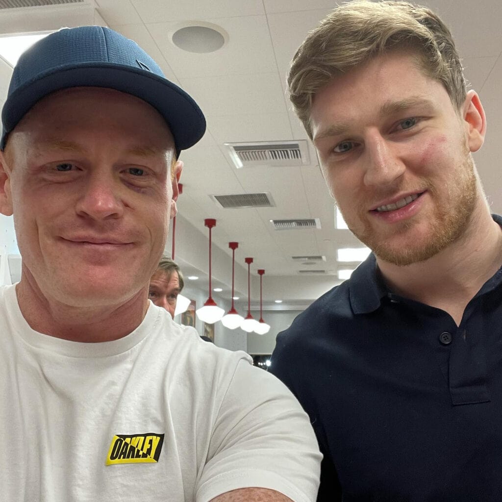 Two men stand close together at In-N-Our, smiling at the camera. One, Nate MacKinnon, wears a navy shirt, the other, Brent, a white Oakley t-shirt and blue cap.