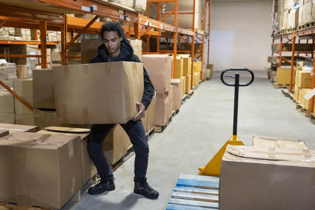 Warehouse worker lifting a large cardboard box using improper posture, highlighting the risks of poor manual handling and the importance of proper training to prevent musculoskeletal injuries.