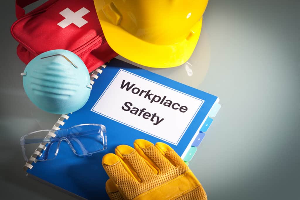 A flat lay showing a blue “Workplace Safety” manual surrounded by a yellow hard hat, light blue respirator mask, clear safety goggles, yellow work gloves and a red first-aid kit.