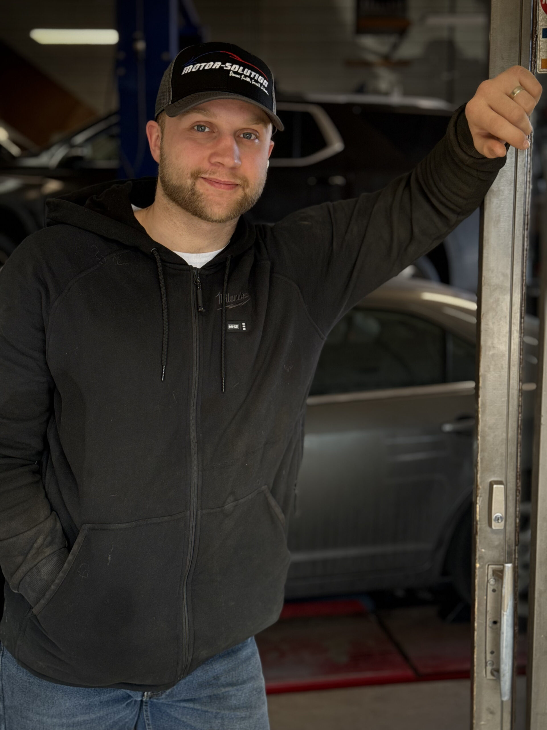 Friendly Motor Solution technician in workshop with cars in background.