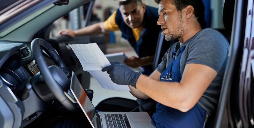Auto repair technician inspecting vehicle inside the car with a laptop and paperwork.
