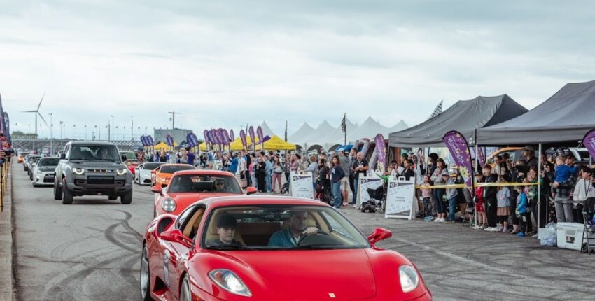 Red sports car driving at a car event with tents and spectators in the background.