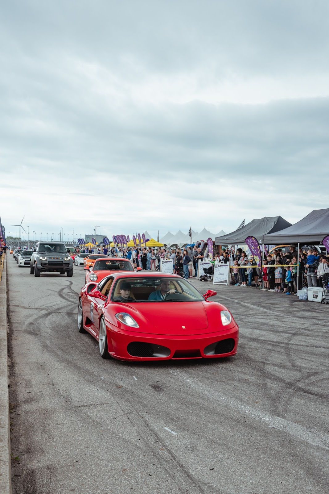 Red sports car driving at a car event with tents and spectators in the background.