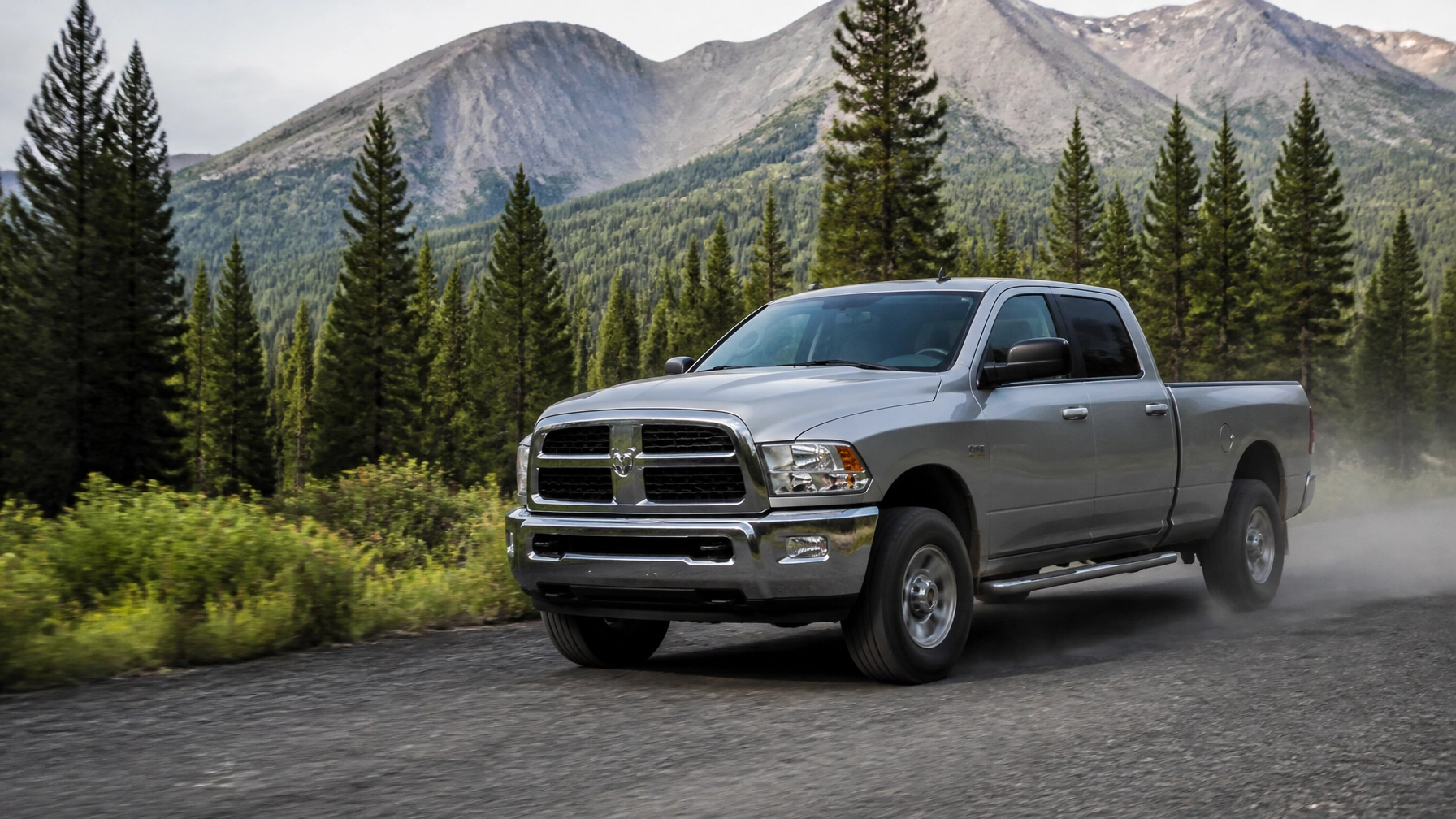 Silver pickup truck driving on mountain road surrounded by pine trees and mountains.