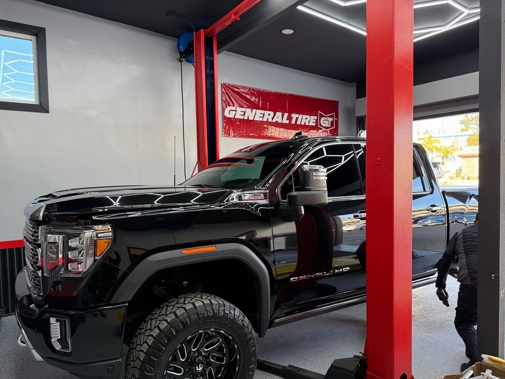 Black heavy-duty truck inside auto repair shop with red lift and General Tire banner.