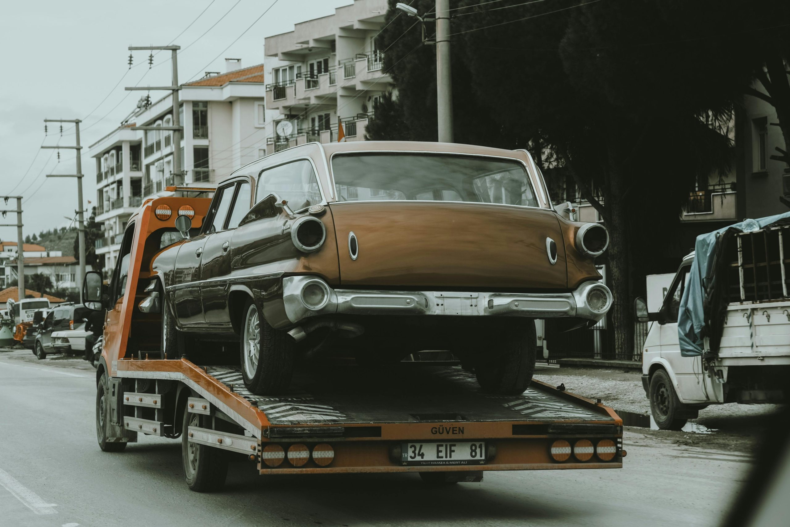 Vintage car being transported on a tow truck in a city street scene.
