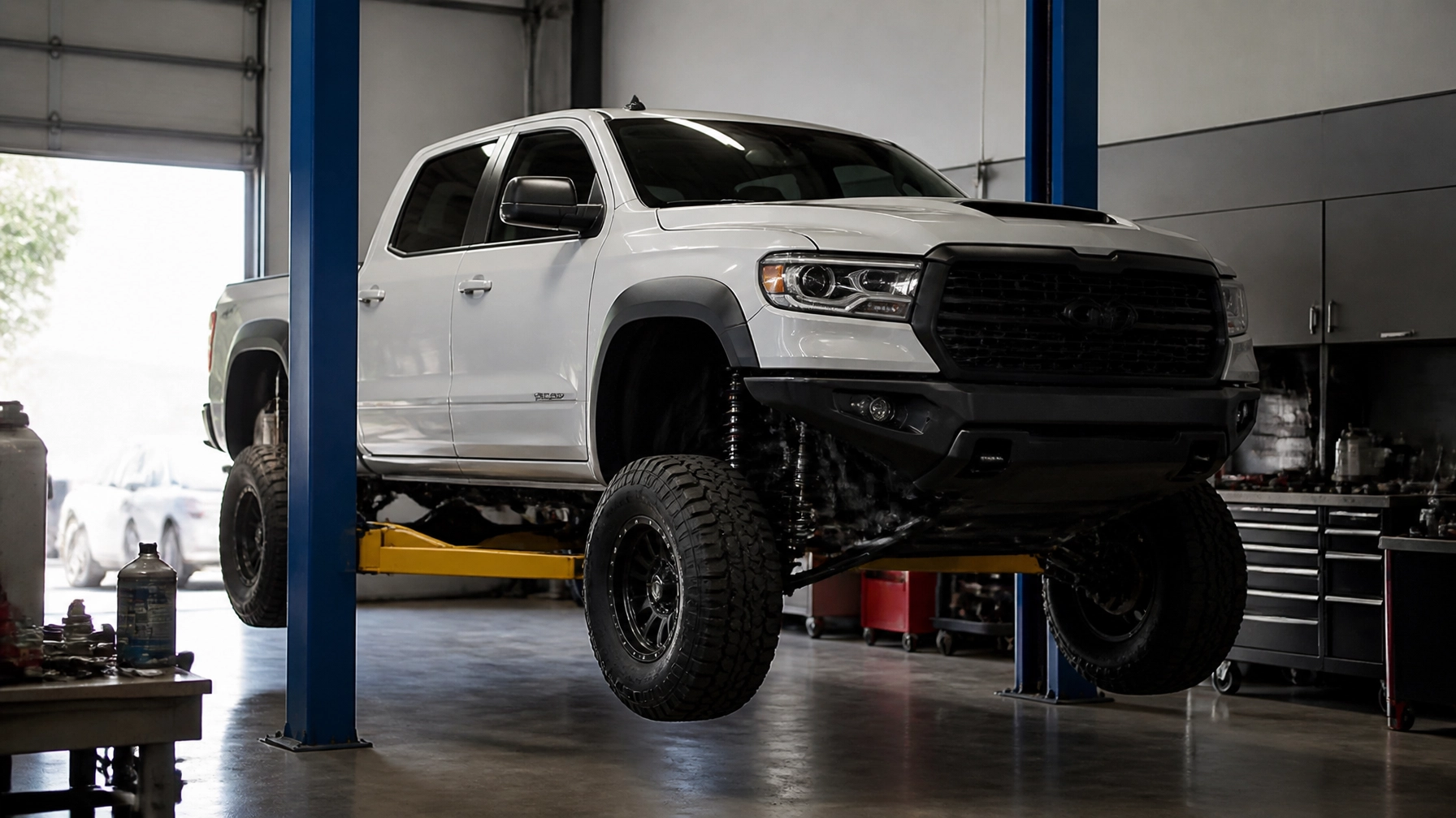 White off-road pickup truck lifted in auto repair shop with tools and equipment.