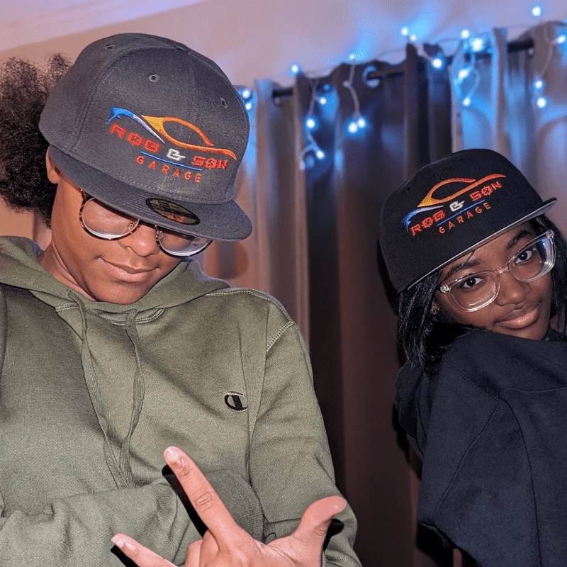 Two young people wearing Rob & Son Garage hats indoors.