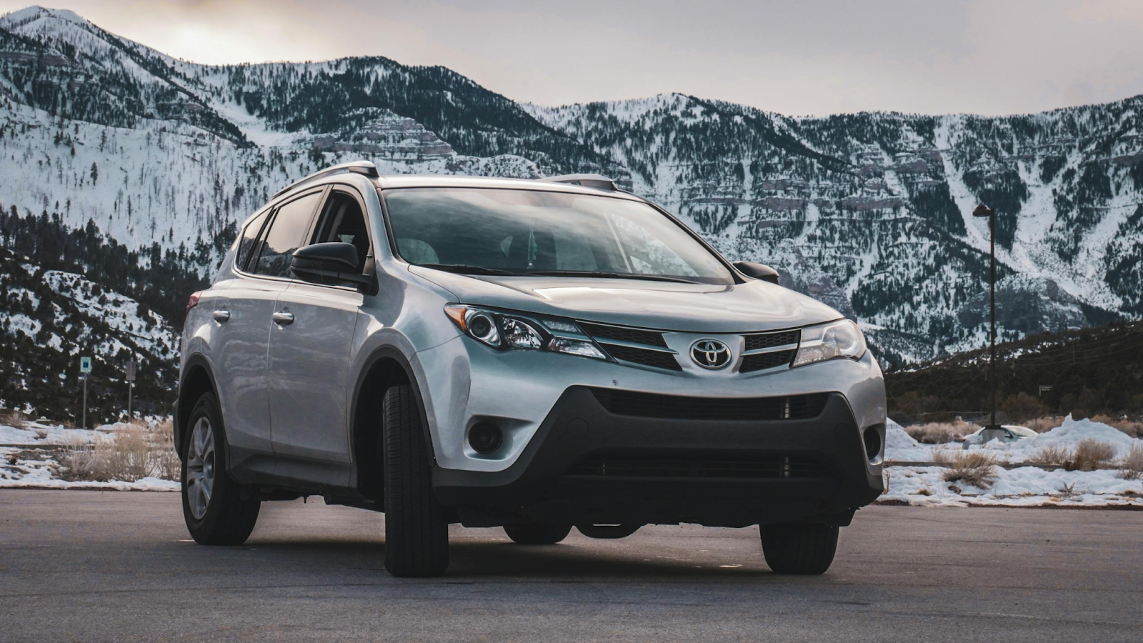Silver SUV parked on mountain road with snow-covered peaks in background.