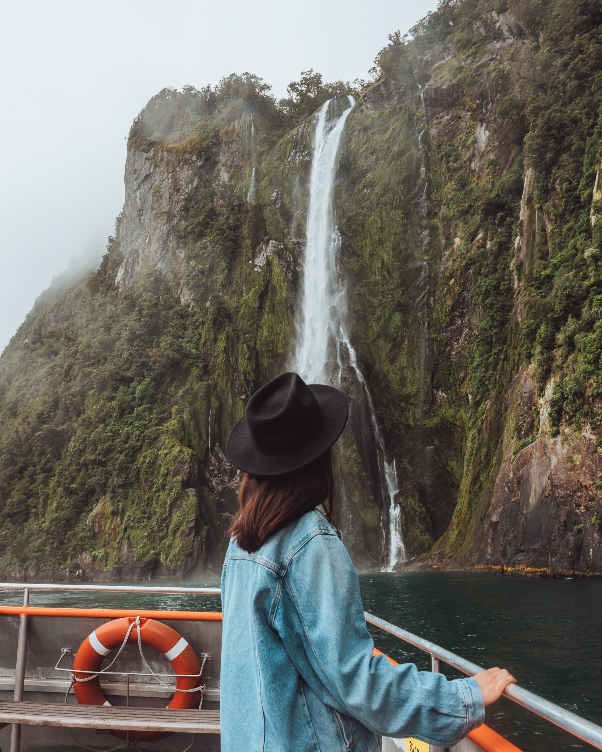 A person in a black hat and denim jacket stands on a boat looking at a tall waterfall cascading down a cliff covered in green vegetation.