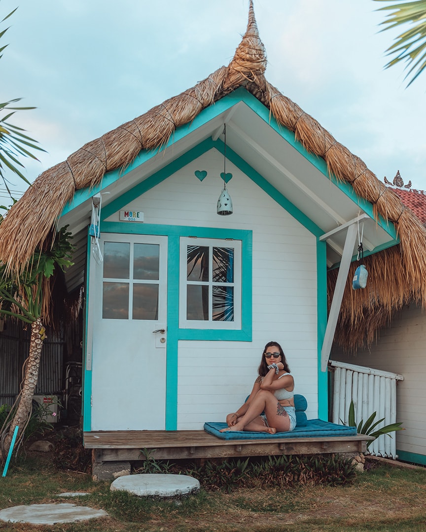 A small white beach hut with turquoise trim and a thatched roof, with a woman sitting cross-legged on the porch, enjoying a quiet moment.