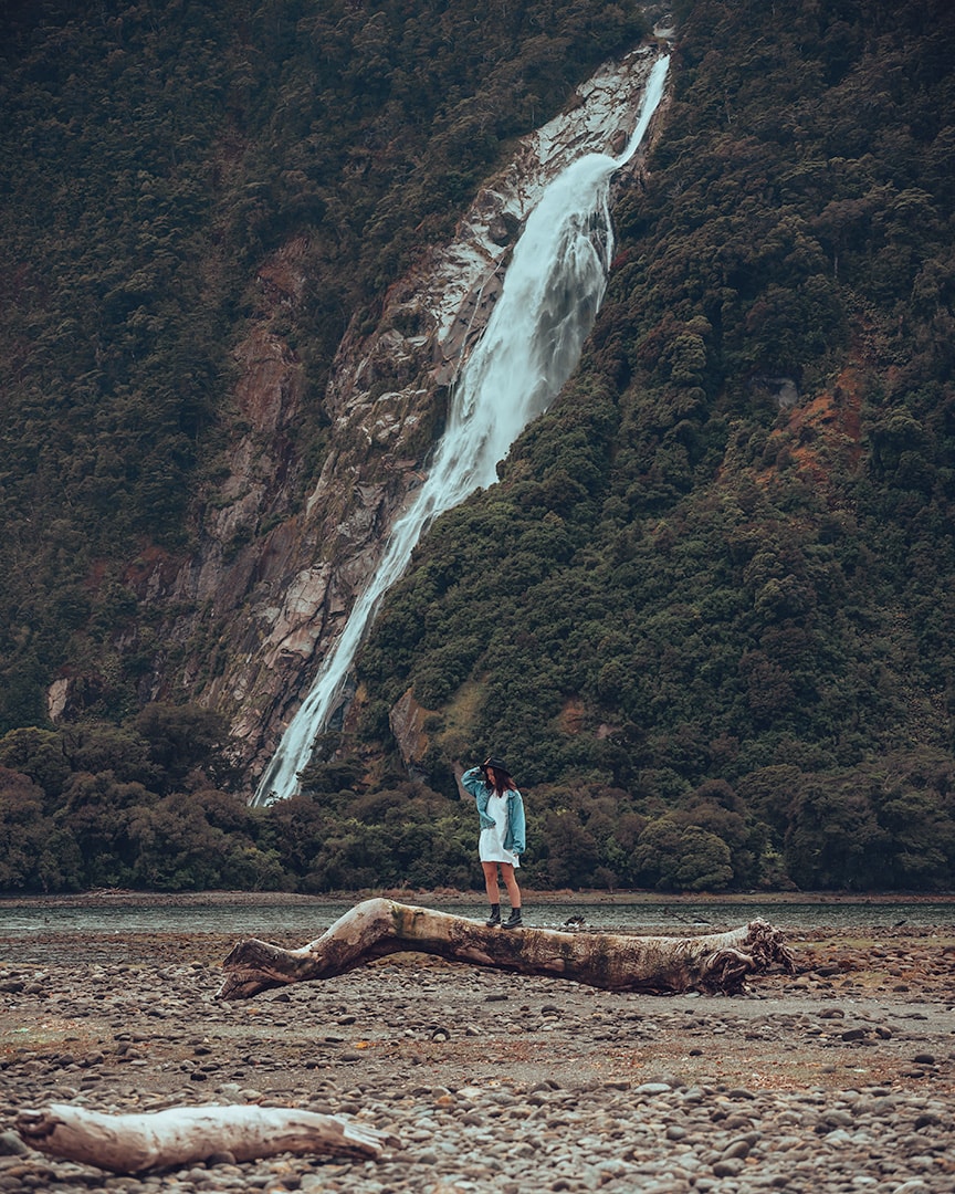 A girl standing on a log in front of a waterfall with lots of lush greenery around it. Milford Sound, New Zealand South Island